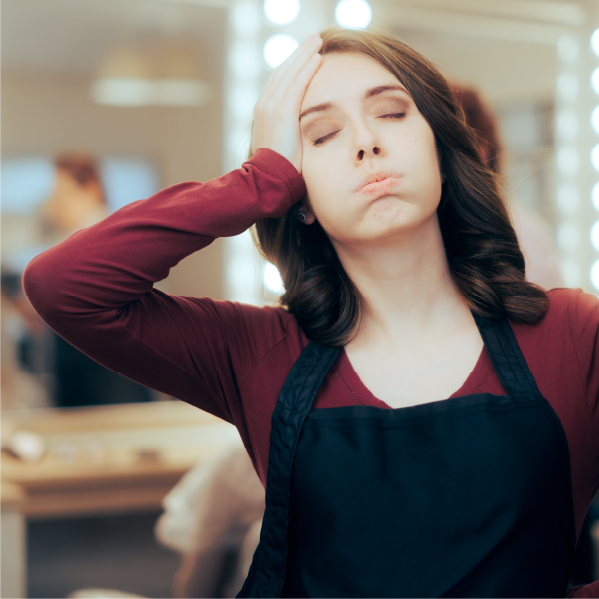Frustrated woman in a beauty salon wearing an apron, resting her forehead on her hand while expressing stress, with soft lighting and mirrors in the background.