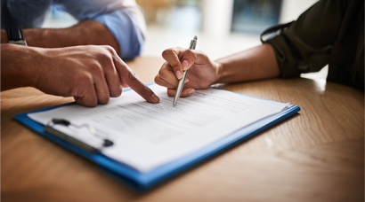 Two hands pointing at a document on a clipboard, with one person holding a pen, indicating a discussion or review of the paperwork.