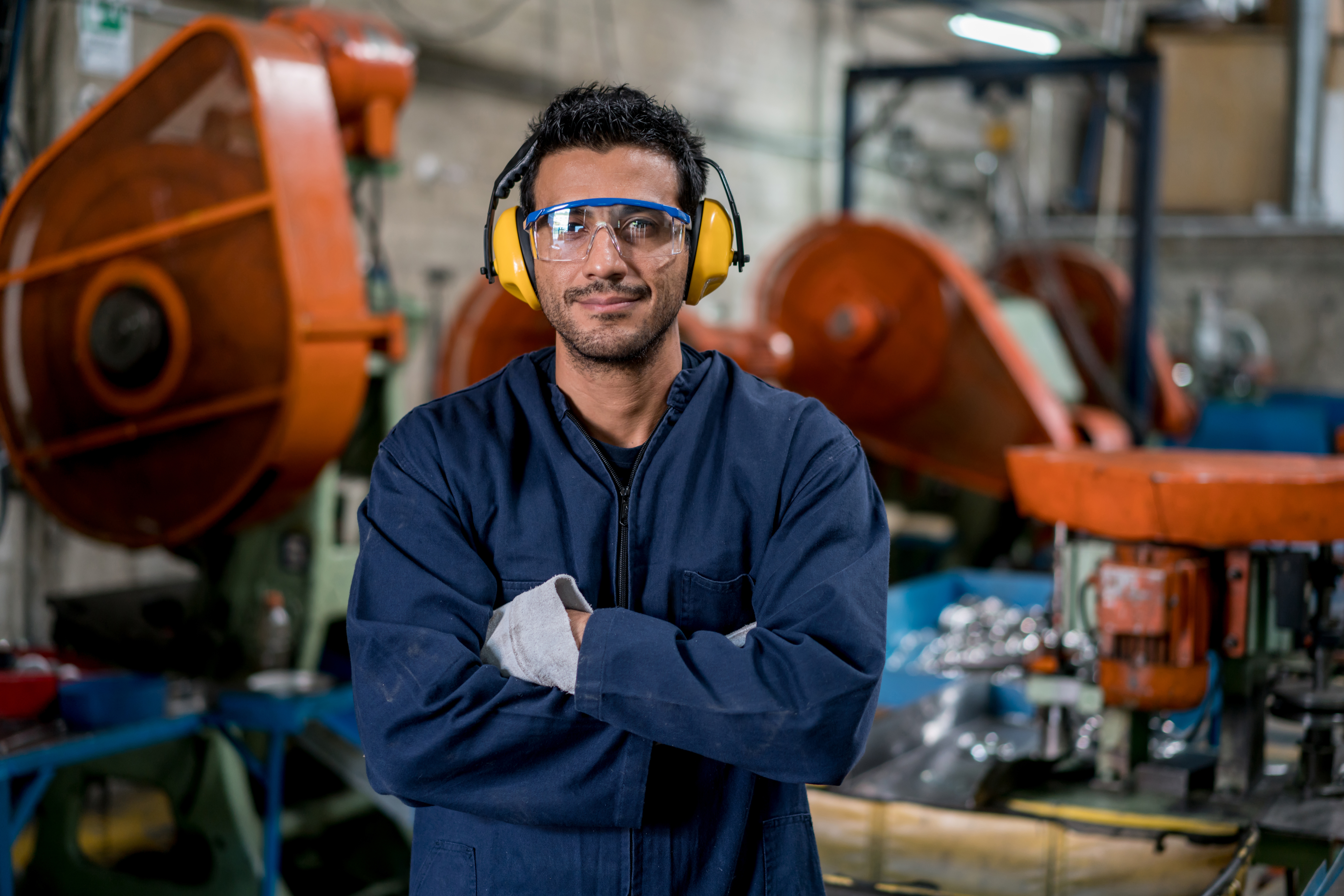 Male factory worker wearing safety glasses and ear protection, standing confidently with arms crossed in an industrial setting filled with machinery and equipment.