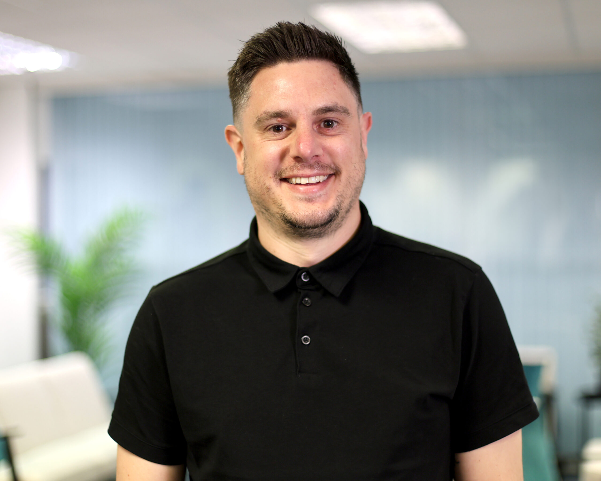 Smiling man in a black polo shirt standing in a modern office environment with plants and soft seating in the background, conveying a professional and approachable atmosphere.