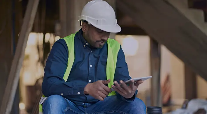 Construction worker in a hard hat and safety vest using a tablet on a job site, focusing on project details.