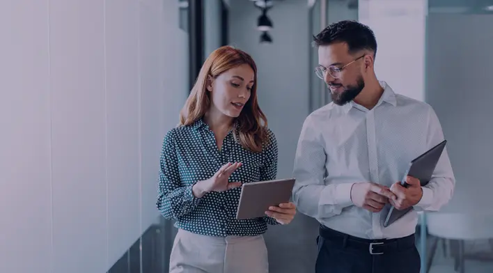 Two professionals discussing a project while reviewing information on a tablet in a modern office hallway. The woman, wearing a polka dot blouse, gestures as she explains something to the man, who holds a tablet and listens attentively.