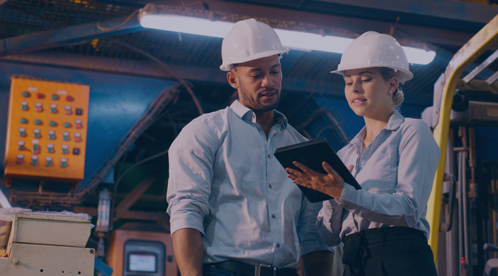 Two construction professionals in hard hats collaborate on a tablet in an industrial setting, surrounded by machinery and control panels, emphasizing teamwork and technology in the workplace.