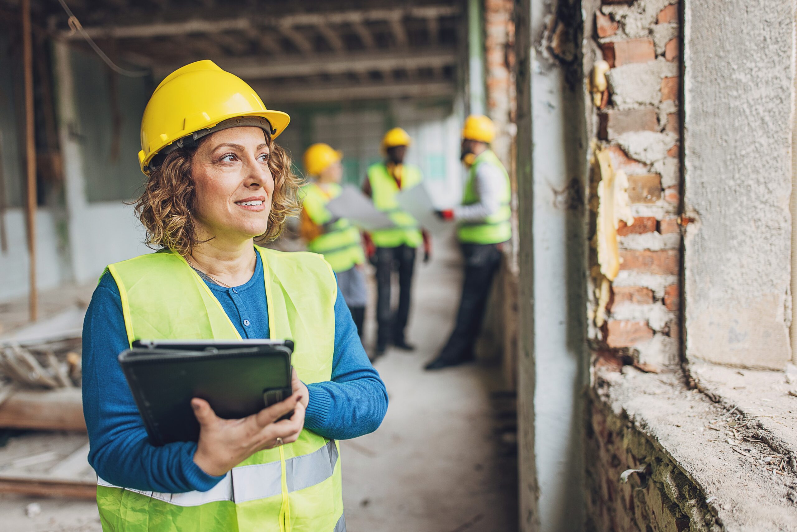 Female construction manager wearing a yellow hard hat and safety vest, holding a tablet, while overseeing a construction site with workers in the background. The image highlights leadership in the construction industry.